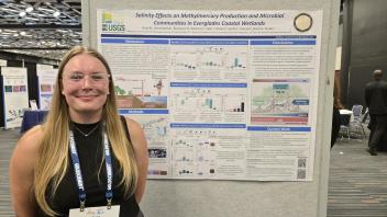 Smiling woman with glasses and conference badge standing by a scientific poster in a hall